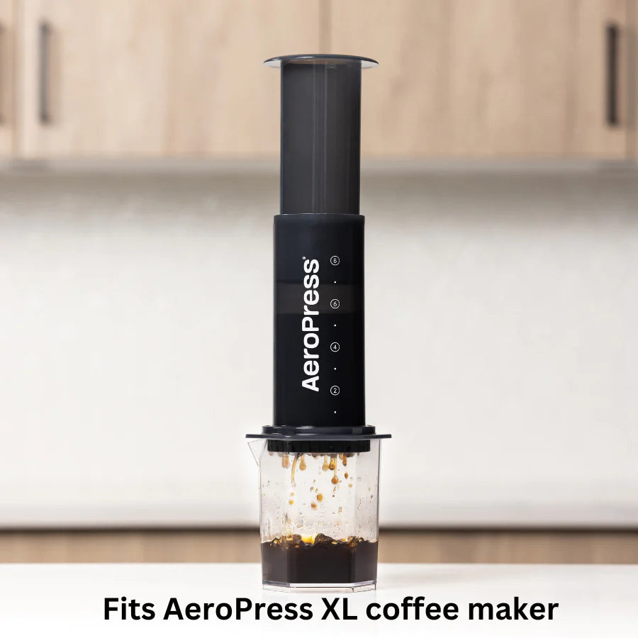 En sort AeroPress kaffemaskine brygger frisk kaffe til en klar kop. Teksten lyder: "Passer til AeroPress XL Filterpapir." En sløret køkkenbord og et skab forstærker den varme stemning. Mærke: Aeropress.
