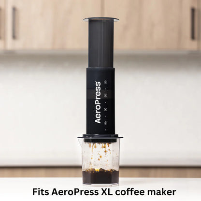 En sort AeroPress kaffemaskine brygger frisk kaffe til en klar kop. Teksten lyder: "Passer til AeroPress XL Filterpapir." En sløret køkkenbord og et skab forstærker den varme stemning. Mærke: Aeropress.