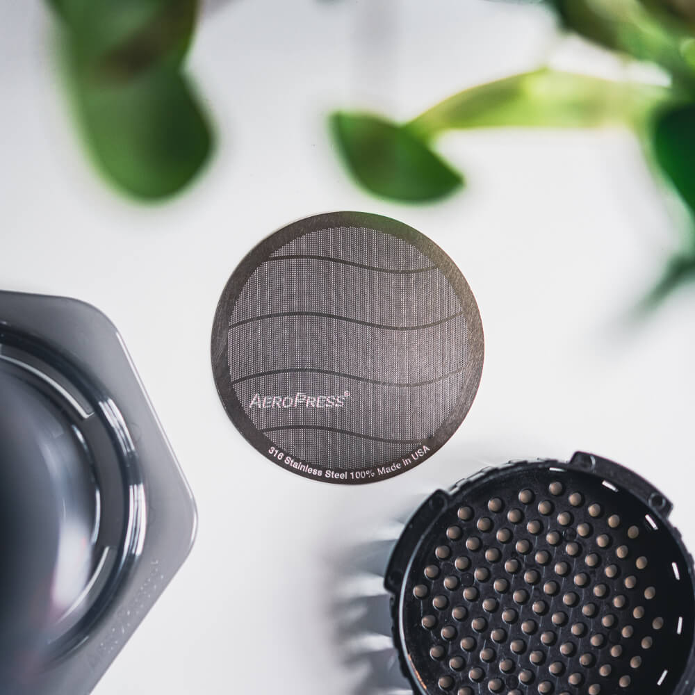 Nærbillede af AeroPress metalfilteret med teksten "Aeropress 3rd Stainless Steel 100% Made in USA", med et bølget rustfrit stål 316 design. Slørede elementer: AeroPress-brygger, perforeret holder, frodige grønne blade i baggrunden.