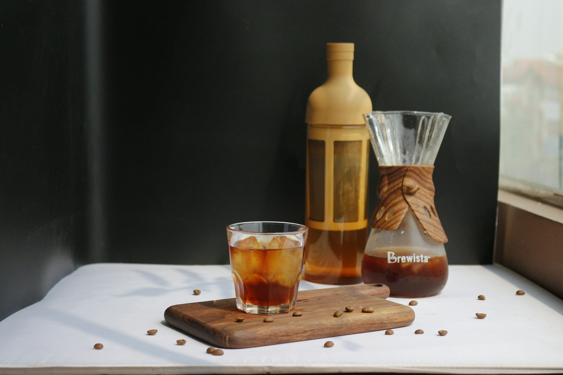 Et glas EverBean Limited ICED Coffee Blend (w) står på et træbræt med Arabica-bønner ved siden af en Brewista-karaffel med cold brew og en brun flaske, alt sammen arrangeret på en hvid overflade mod en mørk baggrund.
