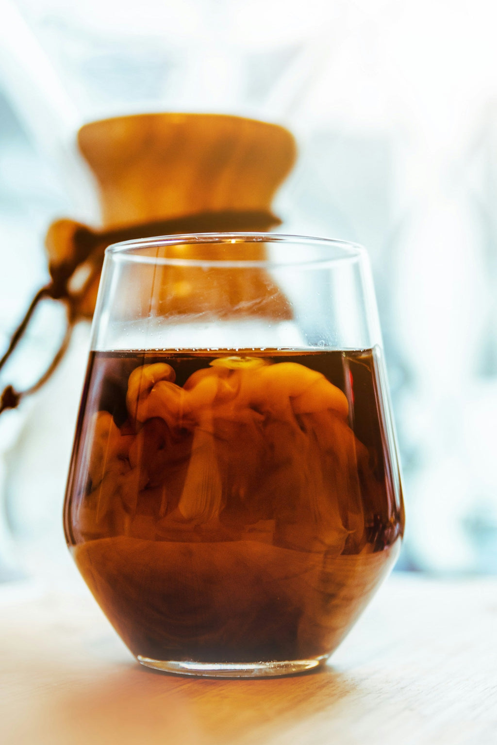 Et nærbillede af et glas fyldt med EverBean Limiteds ICED Coffee Blend (w), hvor mælk hvirvler rundt indeni. En Chemex-kaffemaskine i baggrunden antyder friske Arabica-bønner, badet i klart naturligt lys, hvilket skaber et sprødt og forfriskende billede.