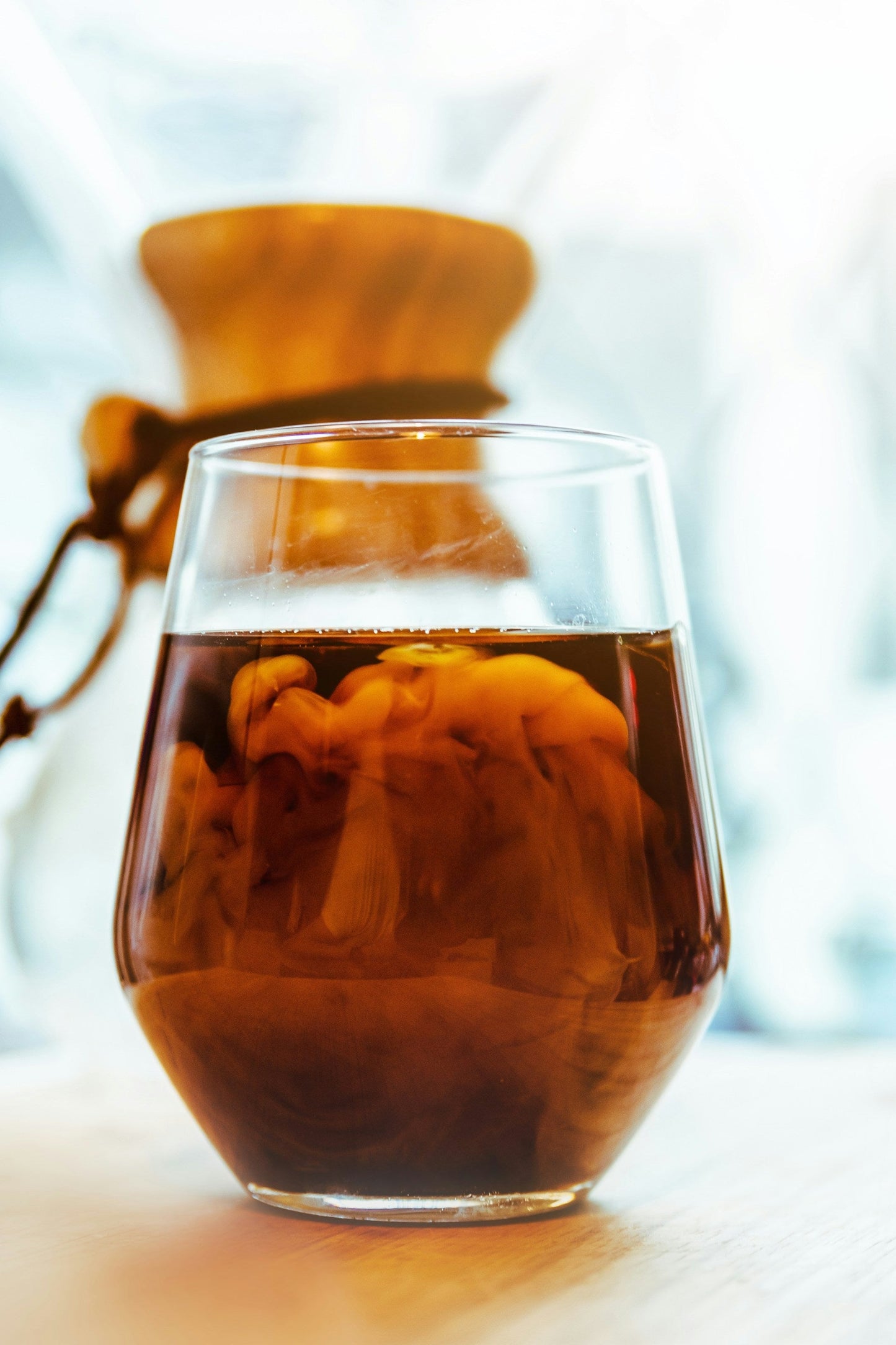 Et glas iced ICED Coffee Blend (b) fra EverBean blandet med mælk hvirvler yndefuldt. I den blødt oplyste baggrund hviler en Chemex kaffemaskine på bordet og fremkalder en fredfyldt morgenstemning forstærket af den rige aroma af Arabica bønner.