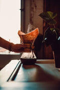 En hånd holder en Chemex-kaffekande af træ fyldt med dampende kaffe på et bord ved siden af en potteplante, mens varmt sollys strømmer ind gennem et vindue i baggrunden.