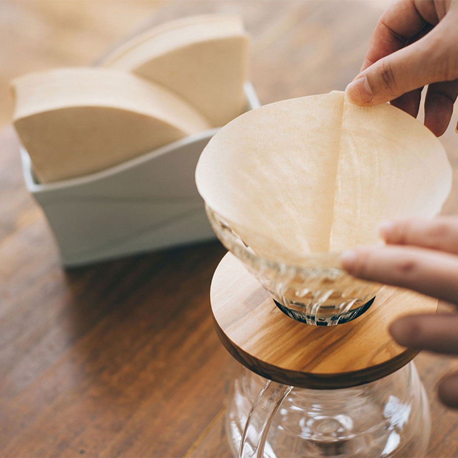 En person arrangerer præcist en Hario, Papir Filter, Størrelse 02 i en kaffedryp af glas på et træstativ og nyder kaffebrygningsritualer. I nærheden hviler en kasse med lignende filtre på det rustikke træbord.