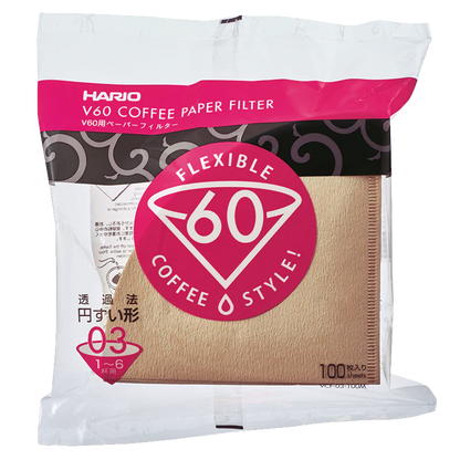 En pakke med 100 Hario Papirfiltre, naturbrune kegleformede kaffefiltre (størrelse 03), biologisk nedbrydelige, i klar plastfolie med pink og hvid "FLEXIBLE V60 COFFEE STYLE!"-etiket. Mærke: Hario.