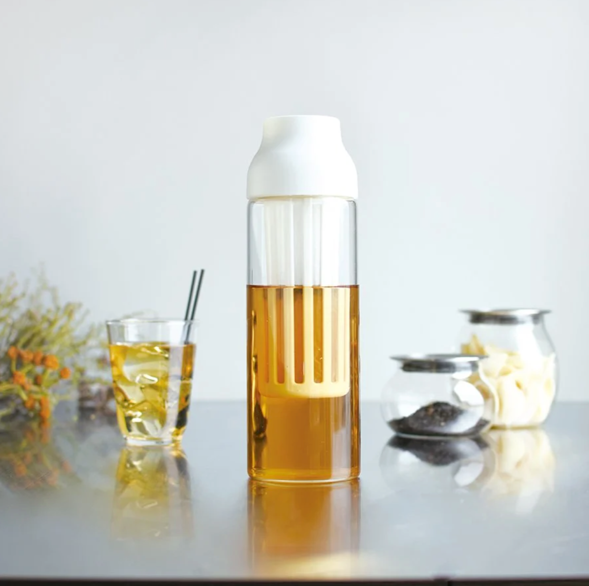 En høj Kinto Capsule Coldbrew Karafel, fyldt med ravfarvet væske, svarende til koldbrygget kaffe, og toppet med et hvidt låg, sidder på en reflekterende overflade. I nærheden er et glas af den samme drink, plus krukker med teblade og hvide genstande. Grønt fremhæver baggrunden.