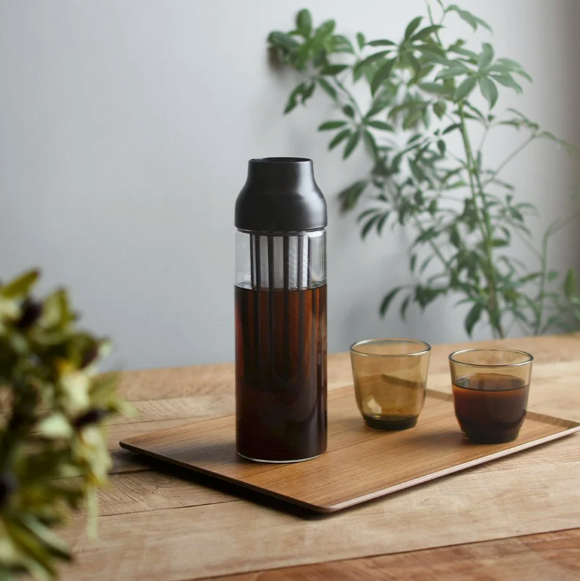 En Kinto, Capsule Coldbrew Karafel sidder på en træbakke med to små brune glaskopper, omgivet af grønne bladplanter for en indbydende og forfriskende atmosfære.