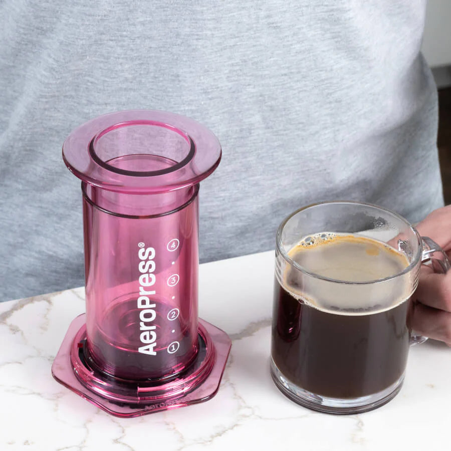 En person i en lysegrå skjorte står ved en bordplade og holder et klart glaskrus fyldt med kaffe ved siden af deres innovative pink AeroPress, Clear Kaffebrygger lavet af Aeropress af slidstærkt Tritan™-materiale.