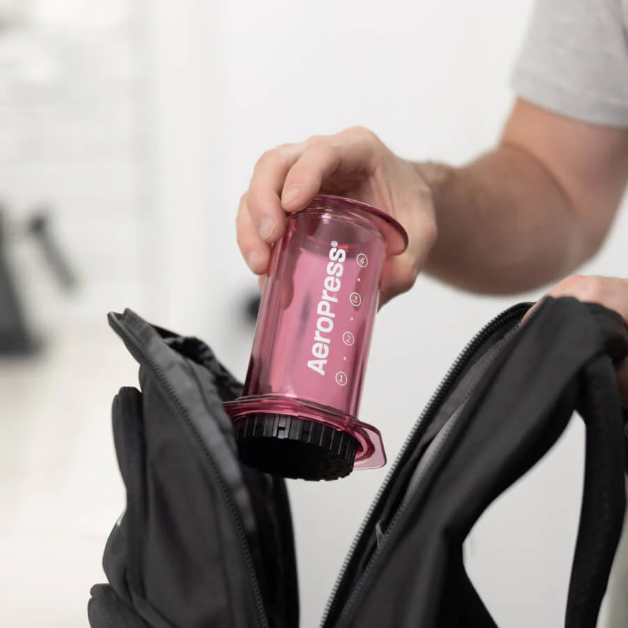 En person pakker forsigtigt deres pink-limited AeroPress Clear Kaffebrygger fra Aeropress, med innovativ bryggeteknologi og holdbar Tritan™, i en slank sort rygsæk.