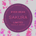 En cirkulær etiket med "Sakura, Limited, Øko, Single Serve" af EverBean Limited i midten. Lyserød tekst på lilla, indrammet af lyserøde akvarelblomster. Dansk/norsk ring nævner Sakura kaffe og 100 % arabica bønner.