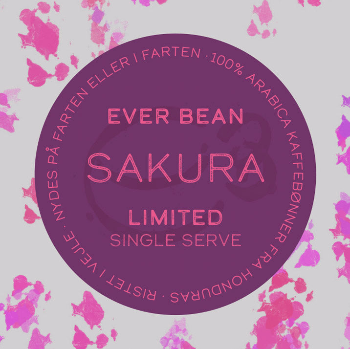 En rund etiket med pink tekst: "Single Serve, Limited, Sakura (øko)" af EverBean Limited på en lilla baggrund. Den omgivende danske tekst omfatter Sakura kaffe og "100% Arabica bønner Honduras", fremhævet med pink akvarelstænk.