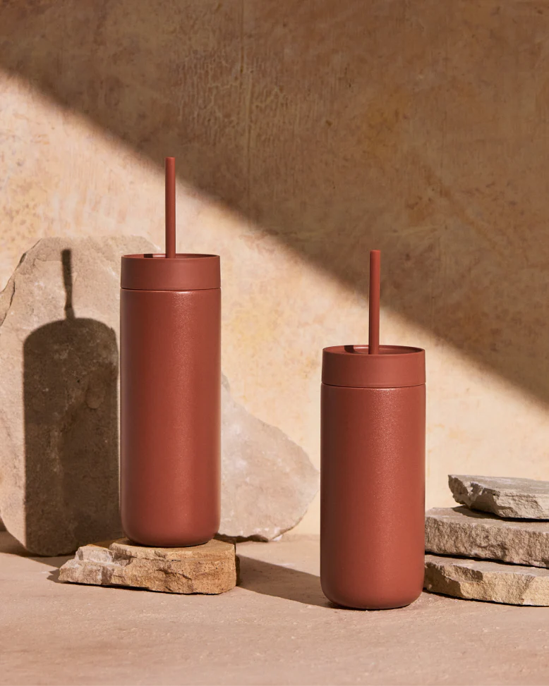 To matte, rustfarvede Fellow Carter Cold Tumbler – 470 ml fra Fellow med tilhørende sugerør står på stenplader foran en beige baggrund. Lys og skygge fremhæver deres design, mens vakuumisoleringen holder drikkevarerne perfekt afkølede.
