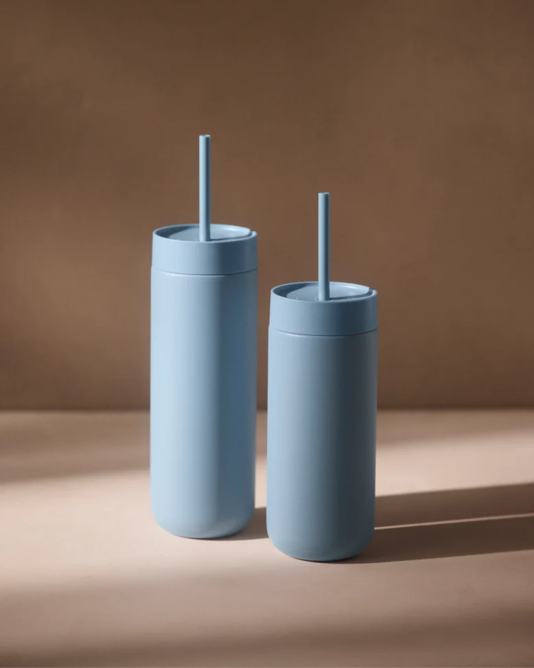 To Fellow Carter Cold Tumbler – 470 ml i mat lyseblå med matchende låg og sugerør står side om side på en neutral brun baggrund, hvor deres vakuumisolering kaster bløde skygger mod højre. Mærke: Fellow.