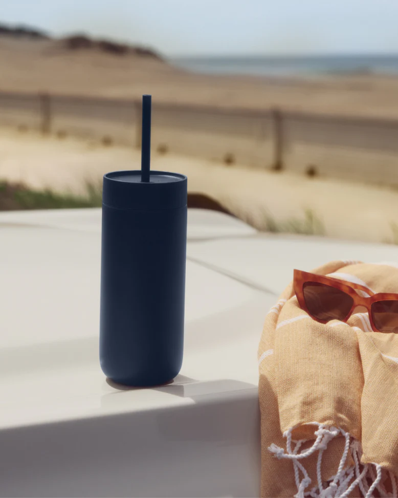 Et Fellow Carter Cold Tumbler-glas – 470 ml fra Fellow i marineblå med et sugerør ligger på en bilhætte ved siden af brune solbriller og et ferskenfarvet tørklæde, mens stranden og havet ses i baggrunden.
