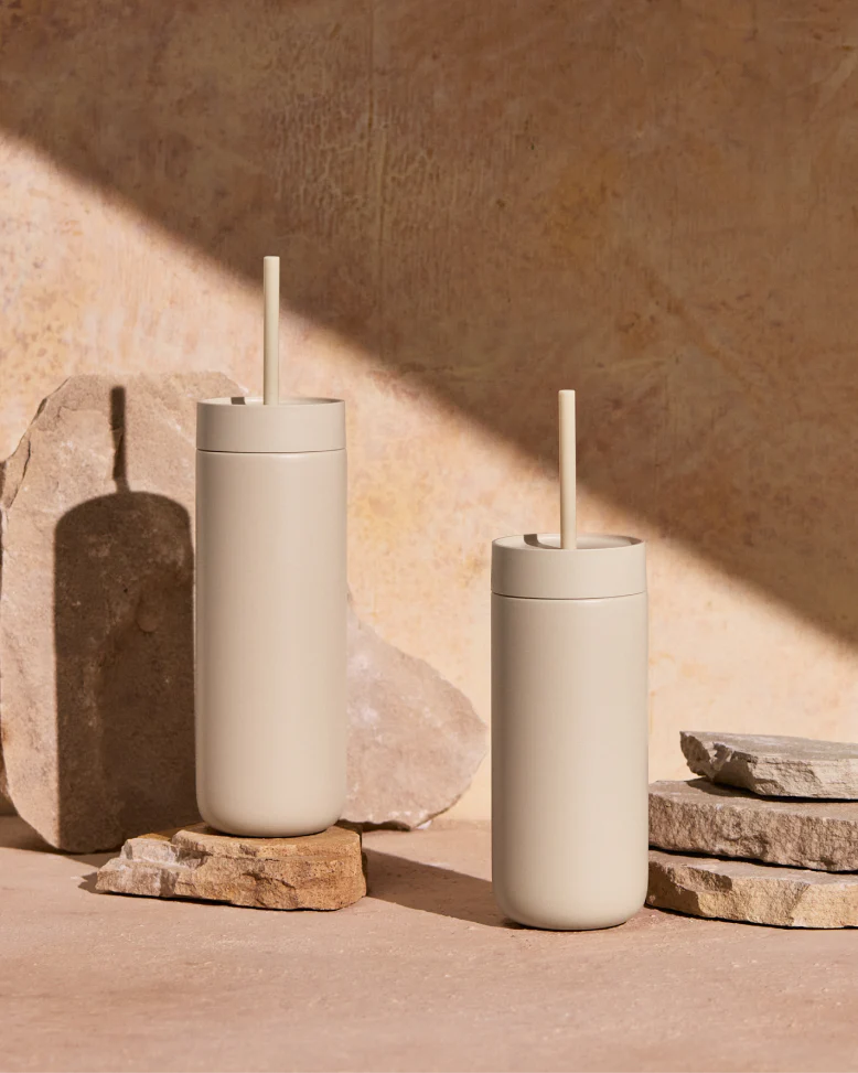 To beige Fellow Carter Cold Tumblers – 470 ml fra Fellow, med sugerør, står på ru stenplader i blødt, naturligt lys. Deres vakuumisolering holder drikkevarerne kolde i timevis.