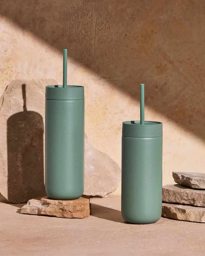 To matgrønne Fellow Carter Cold Tumblers – 470 ml, fra Fellow, er afbildet med sugerør på stenplader foran en neutral baggrund. Den ene tumbler er højere; begge har vakuumisolering og kaster bløde skygger på overfladen.