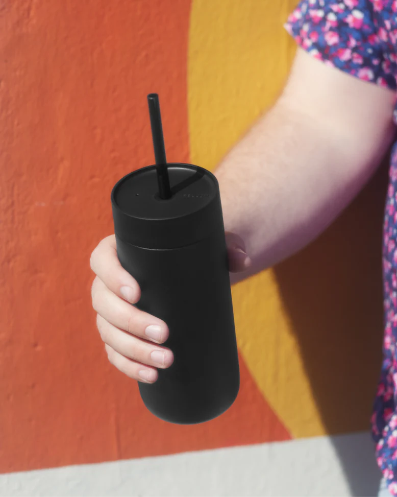 En person i en blomstret skjorte holder en mat sort Fellow Carter Cold Tumbler – 470 ml fra Fellow, komplet med låg, sugerør og vakuumisolering, foran en levende orange og gul væg.