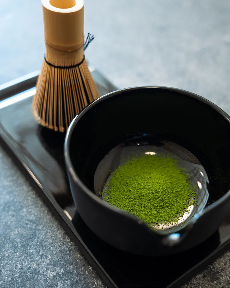 Et Fellow Matcha-sæt – Komplet Matcha-sæt med en sort skål med grønt matcha-pulver står på et sort bakke ved siden af en bambuspiskeris, alt sammen på en grå bordplade. Mærke: Fellow.