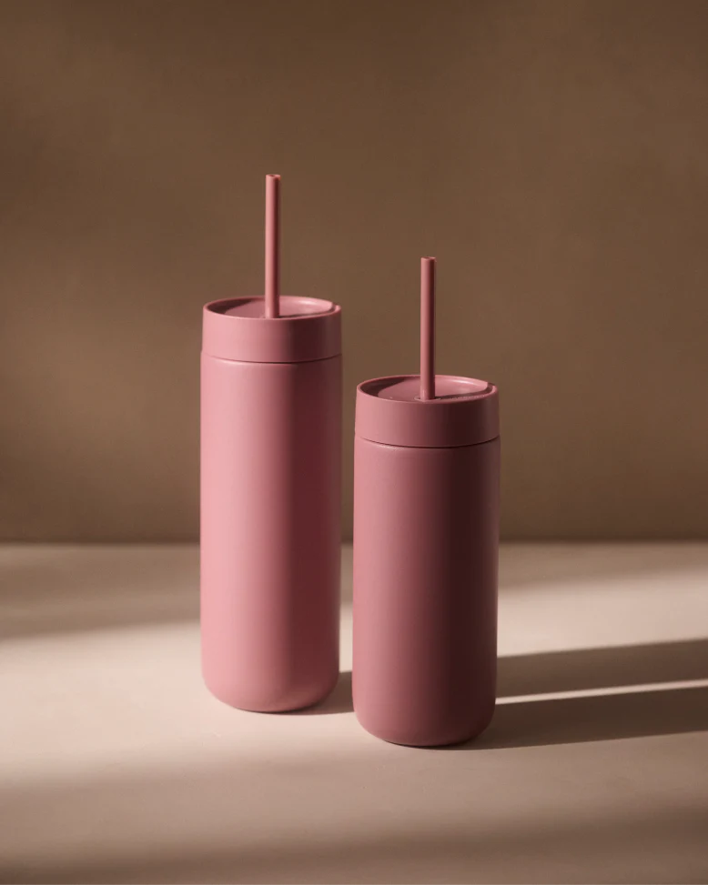 To matrosa Fellow Carter Cold Tumblers (470 ml) fra Fellow med matchende låg og sugerør står side om side på en beige overflade med keramisk belægning, mens det bløde lys kaster milde skygger bag dem.