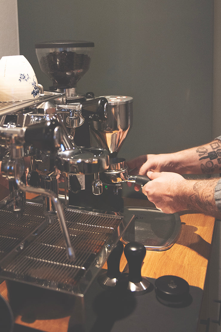 Med tatoverede arme tilbereder en person dygtigt espresso ved hjælp af en kaffemaskine, holder et portafilter midt i bønner og værktøjer som en kværn og tamper på træbordpladen, hvilket sikrer den rige aroma af EverBeans Colombia Supremo fra Espresso smagspakke, mellem fylder luften.