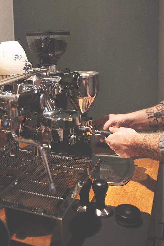 Med tatoverede arme tilbereder en person dygtigt espresso ved hjælp af en kaffemaskine, holder et portafilter midt i bønner og værktøjer som en kværn og tamper på træbordpladen, hvilket sikrer den rige aroma af EverBeans Colombia Supremo fra Espresso smagspakke, mellem fylder luften.